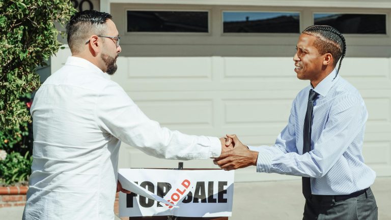 Two men shaking hands in front of a sold house sign, sealing a real estate transaction. Deux hommes se serrant la main devant un panneau "maison vendue", scellant une transaction immobilière.