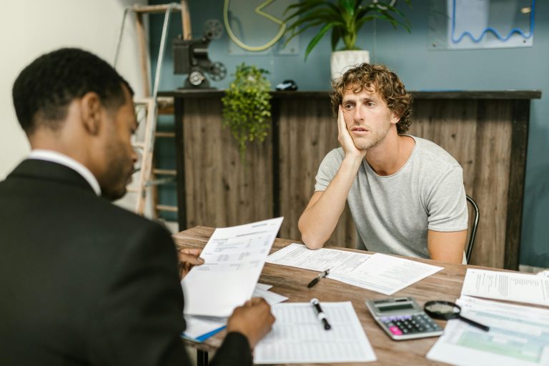 Man in office, appearing stressed during paperwork discussion, sitting opposite professional. Un homme au bureau, paraissant stressé lors d'une discussion sur des documents, assis face à un professionnel.