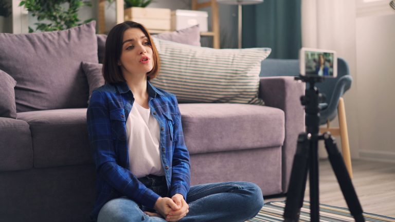 Adult woman recording video indoors on a smartphone, sitting cross-legged on the floor. Femme adulte enregistrant une vidéo à l'intérieur sur un smartphone, assise en tailleur sur le sol.
