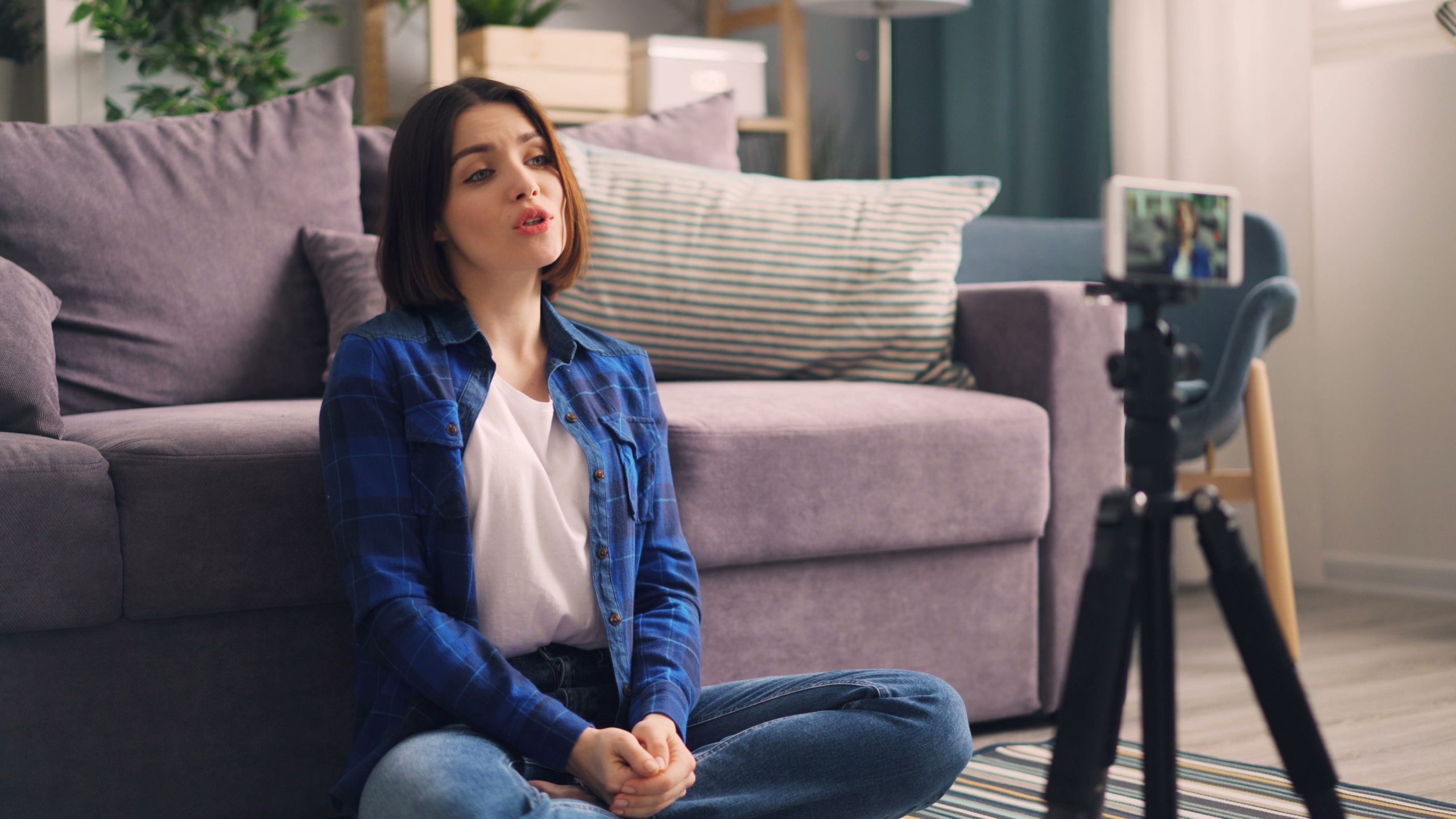 Adult woman recording video indoors on a smartphone, sitting cross-legged on the floor. Femme adulte enregistrant une vidéo à l'intérieur sur un smartphone, assise en tailleur sur le sol.