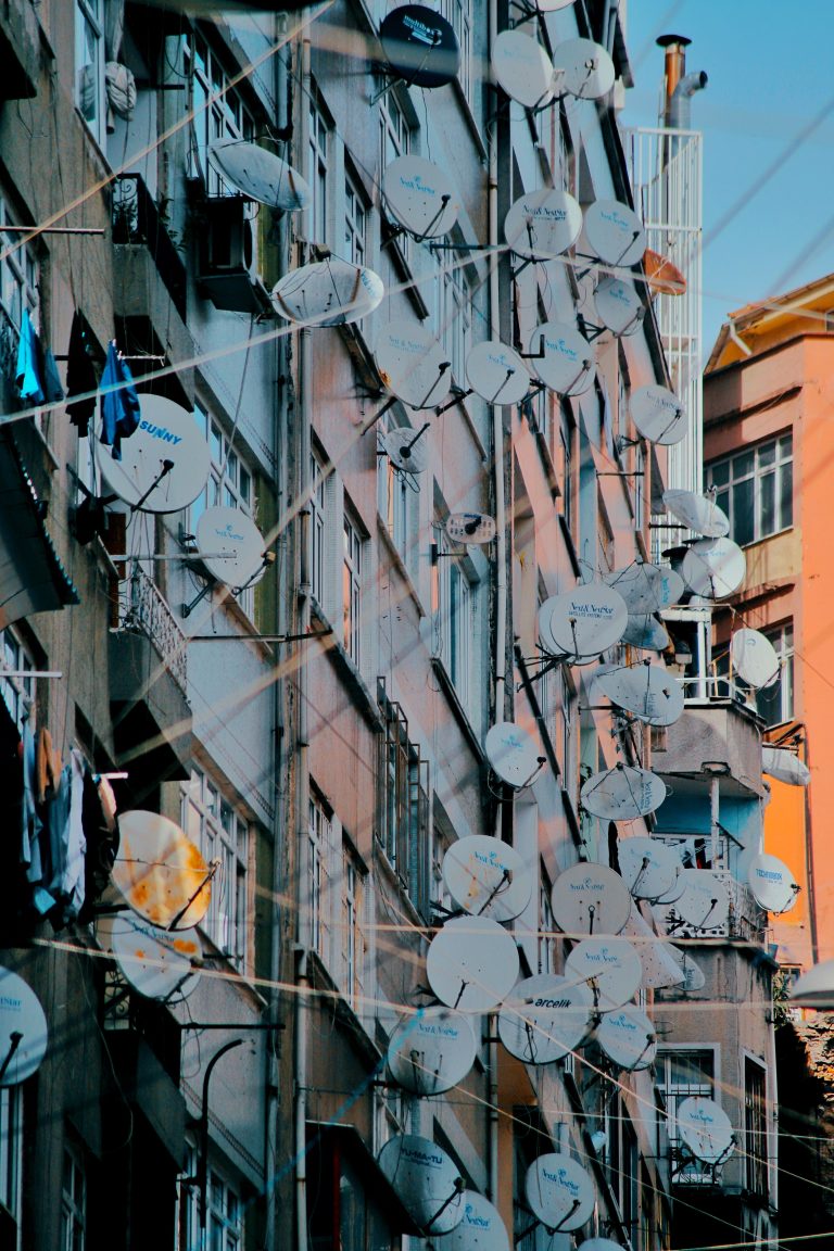A crowded urban building facade adorned with multiple satellite dishes on a sunny day. Une façade de bâtiment urbain bondée ornée de plusieurs antennes paraboliques par une journée ensoleillée.