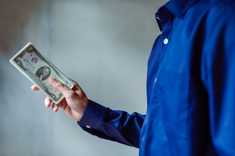 A person in a blue shirt holding a stack of US dollar bills, symbolizing wealth and finance. Une personne en chemise bleue tenant une pile de billets de dollars américains, symbolisant la richesse et les finances.