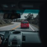 A red car driving on a winding countryside road, viewed from inside another vehicle. Une voiture rouge circulant sur une route sinueuse à la campagne, vue de l'intérieur d'un autre véhicule.