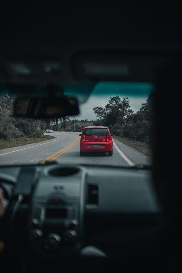 A red car driving on a winding countryside road, viewed from inside another vehicle. Une voiture rouge circulant sur une route sinueuse à la campagne, vue de l'intérieur d'un autre véhicule.