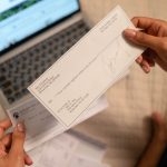 Individual holding a cheque over laptop, signifying online banking or financial transaction. Un individu tenant un chèque au-dessus d'un ordinateur portable, signifiant une opération bancaire en ligne ou une transaction financière.
