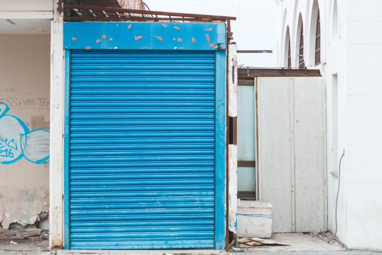 A blue roller shutter on a decayed building facade, featuring graffiti and signs of decay. Un volet roulant bleu sur une façade de bâtiment en décomposition, avec des graffitis et des signes de détérioration.
