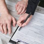 Close-up image of two people signing an insurance policy document on a wooden desk. Image en gros plan de deux personnes signant un document de politique d'assurance sur un bureau en bois.