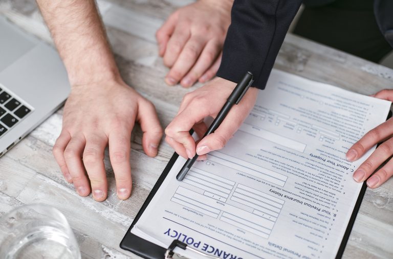 Close-up image of two people signing an insurance policy document on a wooden desk. Image en gros plan de deux personnes signant un document de politique d'assurance sur un bureau en bois.